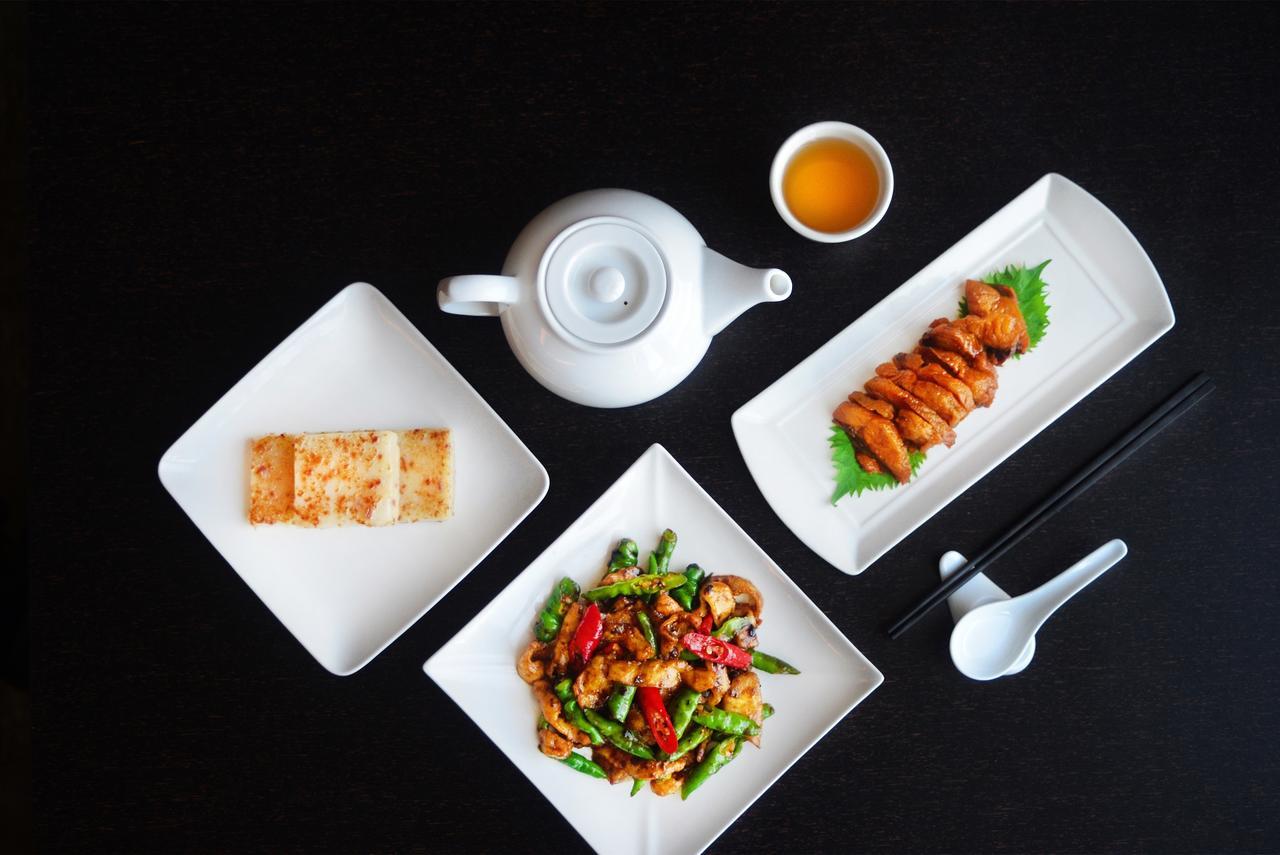 臺北 台北凯达大饭店酒店 外观 照片 The photo features a stylish arrangement of Asian cuisine on a black surface. It includes:

1. **Teapot**: A white teapot positioned with a small cup of tea next to it.
2. **Main Dishes**:
   - A rectangular plate with slices of chicken, likely marin