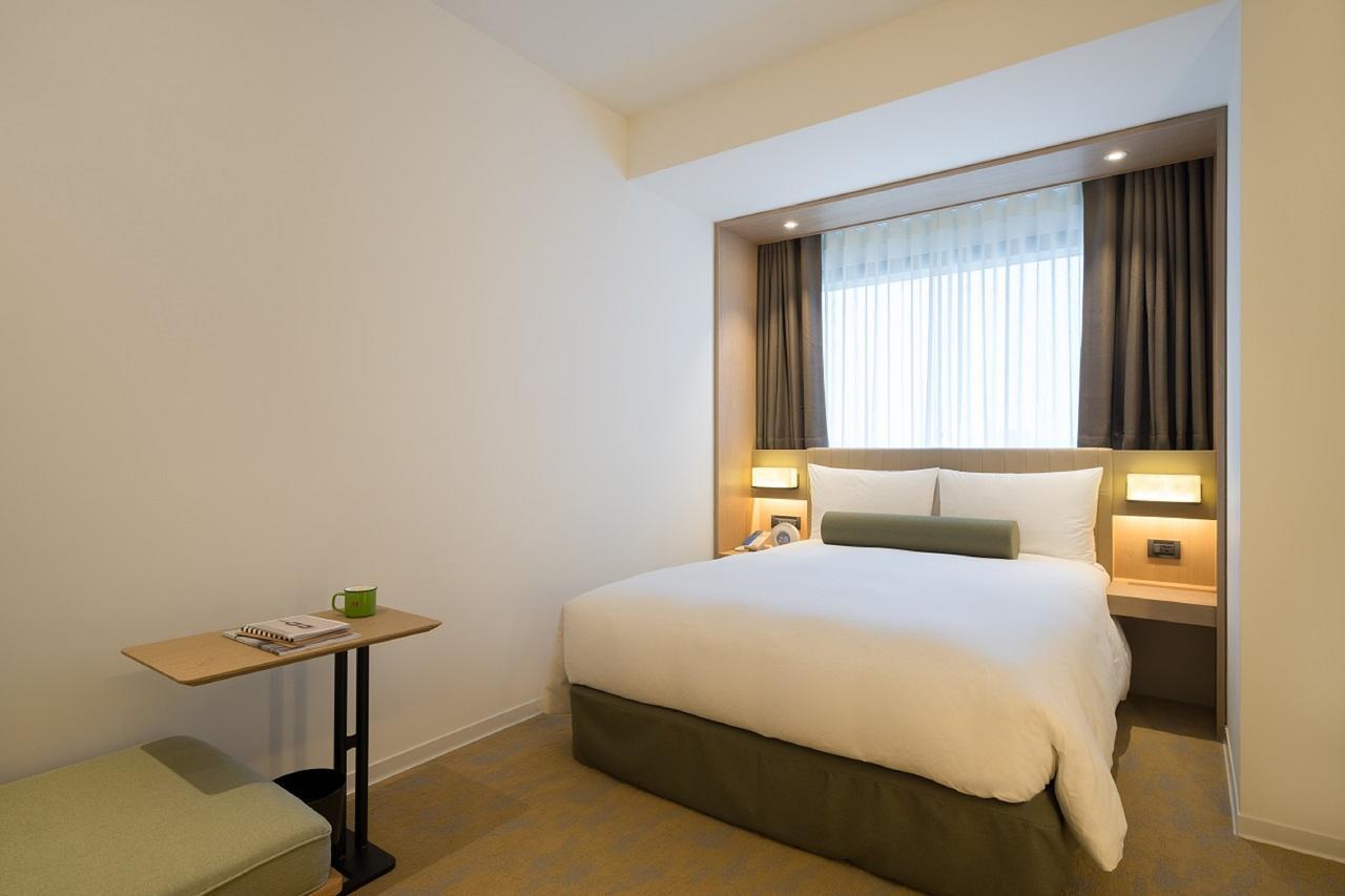 臺北 台北凯达大饭店酒店 外观 照片 The photo shows a modern hotel room featuring a cozy double bed with white bedding and an olive-green base. The room is designed with a minimalist aesthetic, featuring light-colored walls and a small desk with a chair on one side. A window with sheer