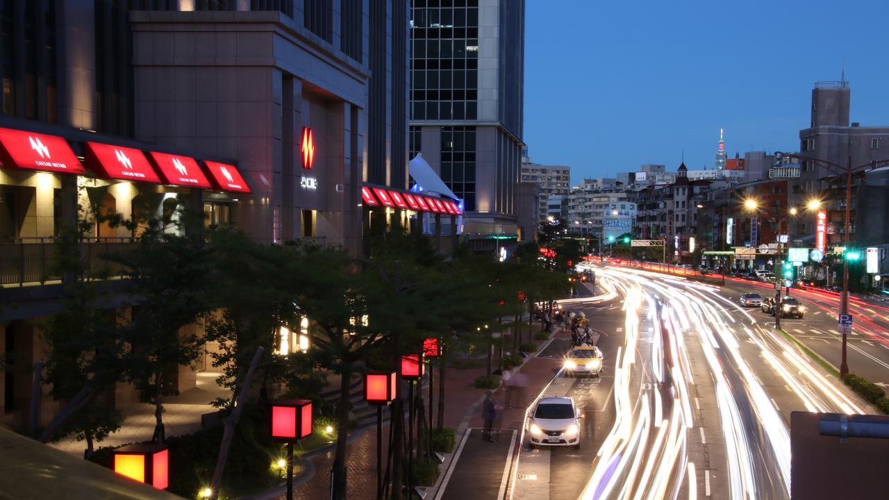 臺北 台北凯达大饭店酒店 外观 照片 The photo shows a city scene during twilight. In the foreground, there is a busy road with streaks of light from moving vehicles, suggesting it's a lively area. On the sides of the street, red lanterns or light fixtures are visible, adding to the amb