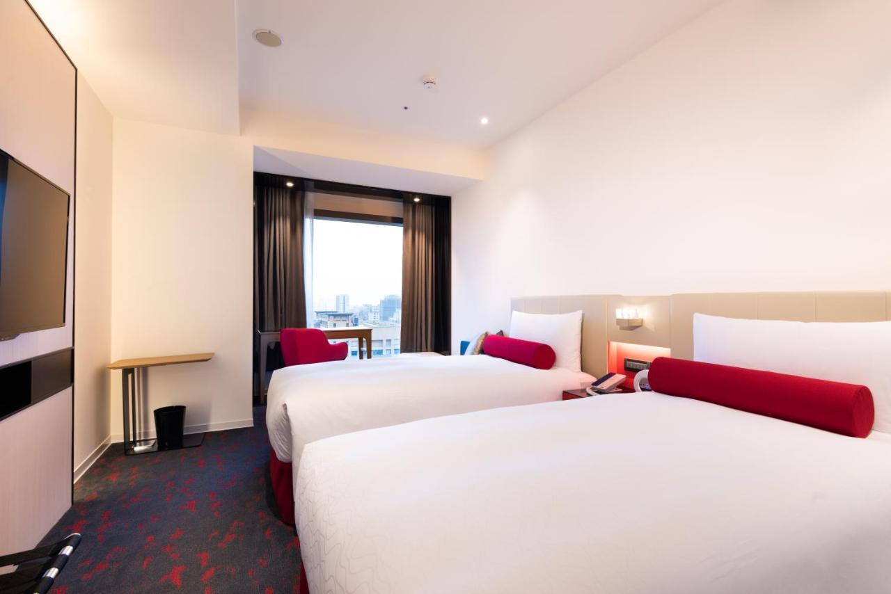 臺北 台北凯达大饭店酒店 外观 照片 The photo shows a modern hotel room featuring two beds with white bedding and red accent pillows. There is a small wooden table next to a window that has dark curtains, allowing for natural light to enter the room. The walls are painted in light colo