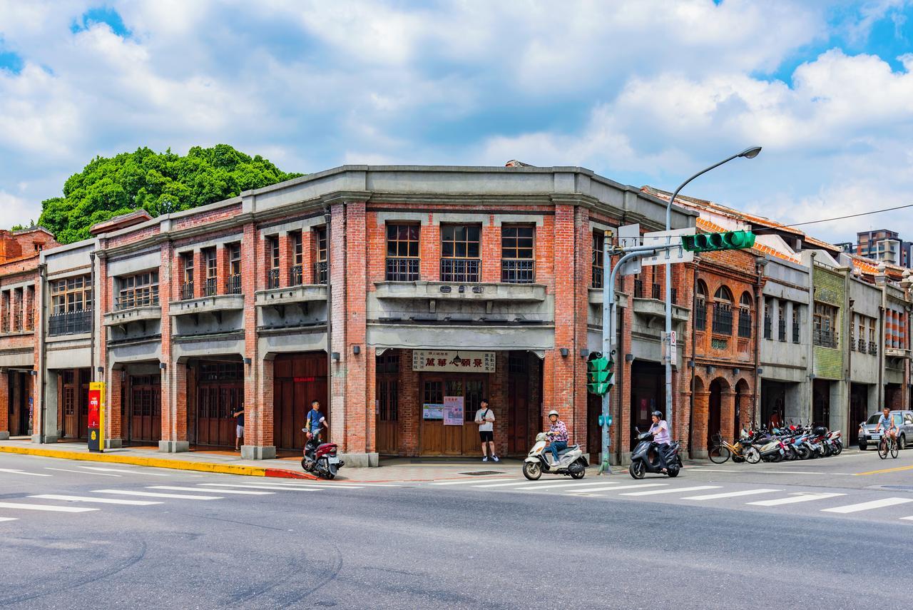 臺北 台北凯达大饭店酒店 外观 照片 The photo shows a street corner featuring a two-story building with a combination of brick and concrete architecture. The building has large windows and a decorative facade, typical of early 20th-century design. In front, there are several people, in