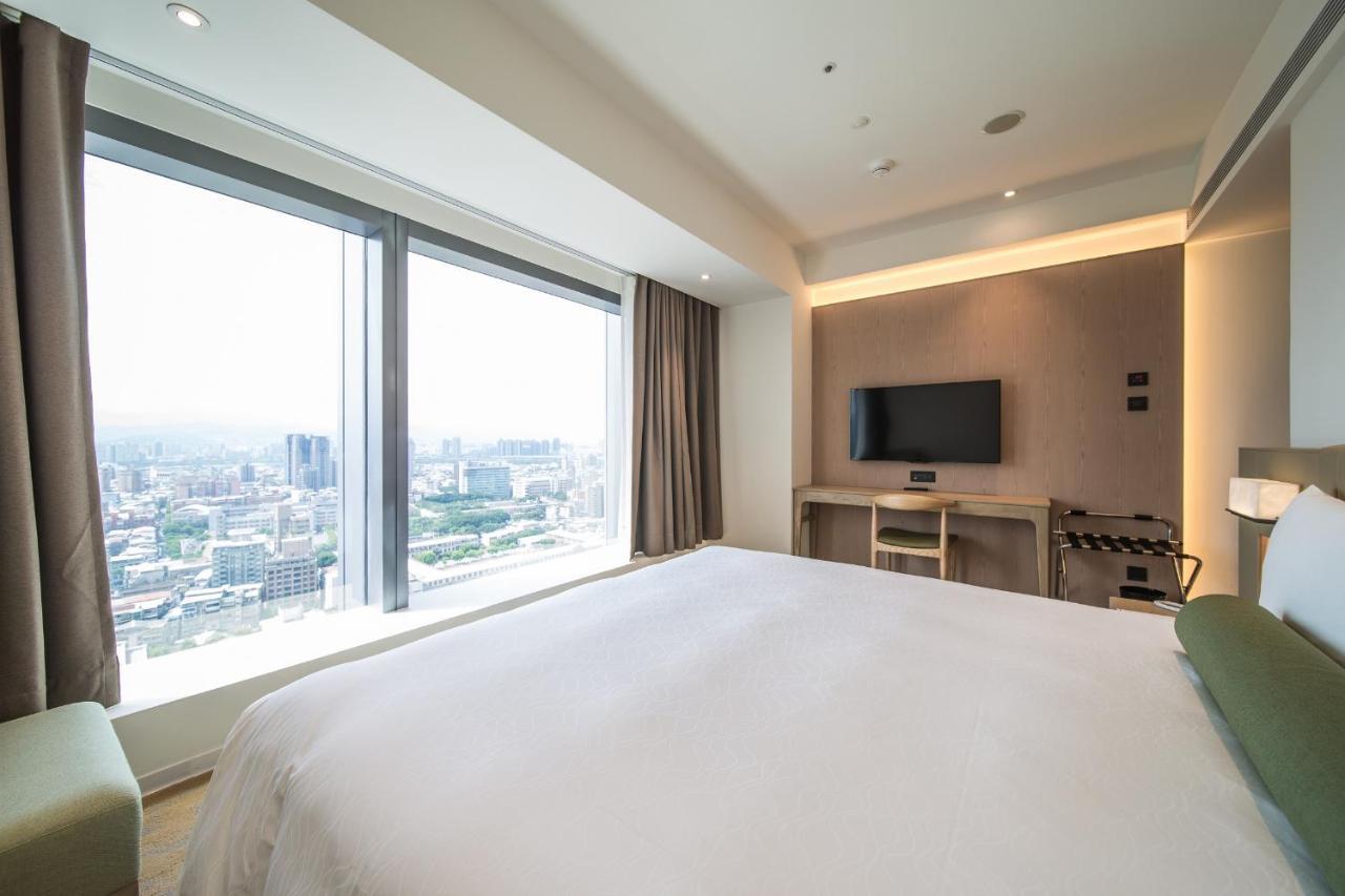 臺北 台北凯达大饭店酒店 外观 照片 The photo shows a modern hotel room featuring a large bed with a white bedspread. There is a large window that offers a view of the city skyline, allowing natural light to fill the space. To the side, there is a flat-screen TV mounted on the wall, an